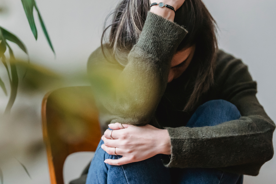 A woman sitting on a wood chair with her elbow on her knees, holding her head. She is dealing with anxiety.