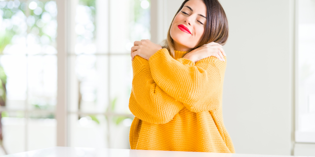 A Caucasian woman wearing an orange-yellow sweater, giving herself a hug. She is demonstrative self-love. She is brunette wearing red lipstick.